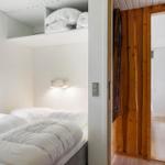 Schlafzimmer mit Bett, Fenster und Aufbewahrung. Holztür führt in einen Flur mit Kleiderständer.