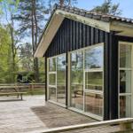 Schwarzes Haus mit Holzterrasse und Glasfront. Umgeben von Bäumen und Gras.