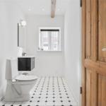 Badezimmer mit Toilette, Waschbecken und Fenster. Holztür und schwarz-weiße Fliesen.