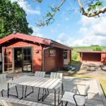Roter Holzhaus mit Terrasse, Grill und Hängeseil. Grün hinter dem Haus.