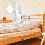 Ein Bett mit Holzgestell, gemustertem Bettbezug und einer roten Decke. Wand mit Tapete und kleinem Fenster.