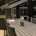 Außenbereich mit Tisch, Stühlen, Blumen und Grill. Hintergrund: Haus und Spielplatz.