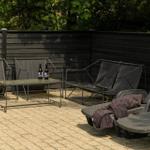 Terrasse mit Liegestühlen, Tisch und zwei Flaschen. Hintergrund: dunkler Zaun und Bäume.