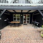 Terrasse mit Tisch, Stühlen, Grill und Pflanzen vor einem Haus mit Balkon.