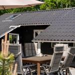 Terrasse mit Tisch, Stühlen und Sonnenschutz vor einem Haus mit dunklem Dach.