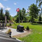 Garten mit Grill, Tisch, Stühlen und dänischer Flagge. Grünfläche und Bäume umgeben den Außenbereich.