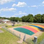 Großer Spielplatz mit buntem Rutschbereich und Schaukelanlage. Hintergrund: Gebäude und Bäume.
