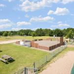 Grüner Außenbereich mit Sandplatz, Picknicktischen und Holzumrandung. Hintergrund: Wald und Gebäude.