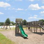 Kinderspielplatz mit Rutsche, Klettergerüst und Schaukel auf Sandfläche unter blauem Himmel.
