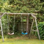 Holzschaukel mit zwei Sitzplätzen im Garten. Hintergrund: Bäume und Sträucher.