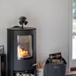 Schwarzer Holzofen mit brennendem Feuer, darunter Holzstapel und daneben Korb mit Brennholz.