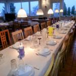 Long table with chairs, tablecloth, dishes, and glasses. Windows with view of trees.