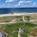 Haus mit Dach aus Stroh, umgeben von Grasland und Strand. Im Hintergrund sind Windräder und das Meer.