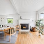 Wohnzimmer mit Kamin, Sofa, Stühlen und Holzboden. Große Fenster und Pflanzen.