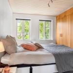 Schlafzimmer mit Bett, Nachttisch, Lampe und Holzschrank. Zwei Fenster zeigen grüne Bäume.