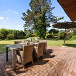 Terrasse mit Tisch, Stühlen und Sonnenschirm. Grünfläche und Baum im Hintergrund.