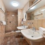 Badezimmer mit Dusche, zwei Waschbecken und Toilette. Wände mit Marmorfliesen verkleidet.