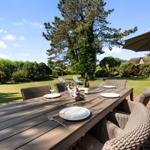 Garten mit Esstisch, Stühlen und Sonnenschirm. Grünflächen und Bäume im Hintergrund.