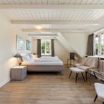 Schlafzimmer mit Doppelbett, Sitzgelegenheiten und großen Fenstern. Holzboden und weiße Wände.