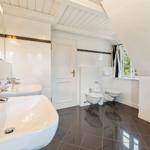 Badezimmer mit zwei Waschbecken, Toilette und Dusche. Weiße Wände und dunkler Boden.