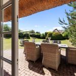Terrasse mit Esstisch und Stühlen unter strohgedecktem Dach. Blick auf grünen Garten und Himmel.