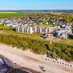 Strand mit Dünen, modernen Gebäuden und grünen Feldern im Hintergrund.