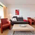 Wohnzimmer mit Sofa, zwei roten Sesseln und Esstisch. Große Fenster und Wandbild.