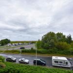 Aussicht auf Parkplatz mit Autos und Wohnwagen, umgeben von Grünflächen und Bäumen unter bewölktem Himmel.