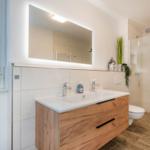 Badezimmer mit Waschtisch, Dusche und Fenster. Holzunterschrank und beleuchtetes Spiegel.