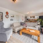 Wohnzimmer mit Sofa, Sessel und Holztisch. Küche im Hintergrund.