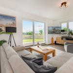 Wohnzimmer mit Sofa, Couchtisch und TV. Große Fenster mit Blick auf Grünfläche.