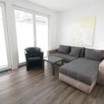 Wohnzimmer mit Sofa, Sessel und Glas-Tisch. Große Fenster und Holzboden.