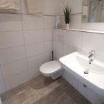 Badezimmer mit Toilette, Waschbecken und Spiegel. Holzoptik Boden. Handtücher und Pflanze.