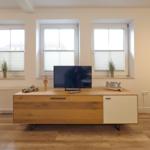 Wohnzimmer mit Holzregal, Fernseher und drei Fenstern mit Jalousien.