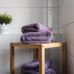 Dusche mit Holzregal, lila Handtücher und Pflanze auf weißer Fliesenwand.