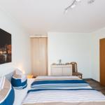 Schlafzimmer mit Bett, Kleiderschrank und Wandbild. Tür und Stuhl sind sichtbar.