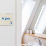 Schild mit Namen La-Trine an der Wand. Im Hintergrund ist ein Badezimmer mit Waschbecken und Spiegel sichtbar.
