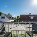 Terrasse mit Tisch und zwei Stühlen. Blick auf Wohnhäuser und Bäume unter blauem Himmel.