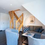 Wohnzimmer mit Sofa, zwei Sesseln und Holztreppe zur oberen Etage.