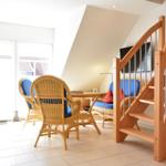 Wohnbereich mit Rattanmöbeln, Holztreppe und TV. Große Fenster mit Blick nach draußen.