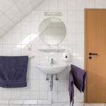 Badezimmer mit Waschbecken, Spiegel und Holztür. Dachneigung und Handtücher sichtbar.