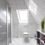 Badezimmer mit Dusche, Toilette und Dachfenster. Weiße Fliesen und Handtuchregal.