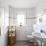 Badezimmer mit Toilette, Waschbecken und Heizung. Holzregal und Fenster mit Pflanze.