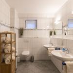 Badezimmer mit Dusche, Waschbecken und Toilette. Holzregal mit Handtüchern.