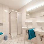 Badezimmer mit Dusche, zwei Waschbecken und Toilette. Blaue Handtücher und Spiegelschrank.