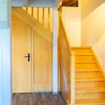 Holztreppe mit Geländer führt zur oberen Etage. Holztür und weiße Ziegelwand sind sichtbar.