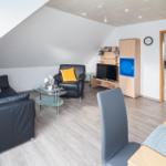 Wohnraum mit Sofa, Sessel, Fernseher und Küchenbereich im Hintergrund.