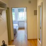 Flur mit Kleiderhaken, Spiegel und Bild an der Wand. Im Hintergrund ist ein Schlafzimmer mit Bett und Fenster.
