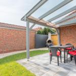 Terrasse mit Tisch, Stühlen und Grill unter Pavillon. Grünfläche und Mauer im Hintergrund.