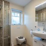 Badezimmer mit Dusche, Waschbecken und Toilette. Fenster sorgt für Tageslicht.